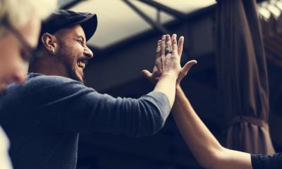Man in a grey sweater and flat cap greeting another person with a high-five